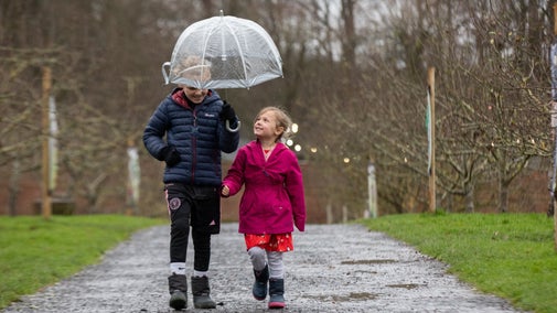 Two children walking along a gravel path smiling underneath a clear umbrella.